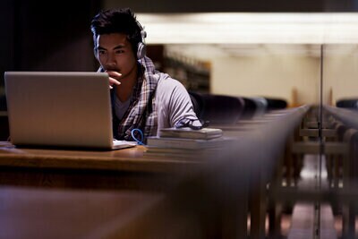 Imagen de un joven con auriculares, sentado frente a una computadora, con libros al lado, en un entorno similar a una biblioteca.
